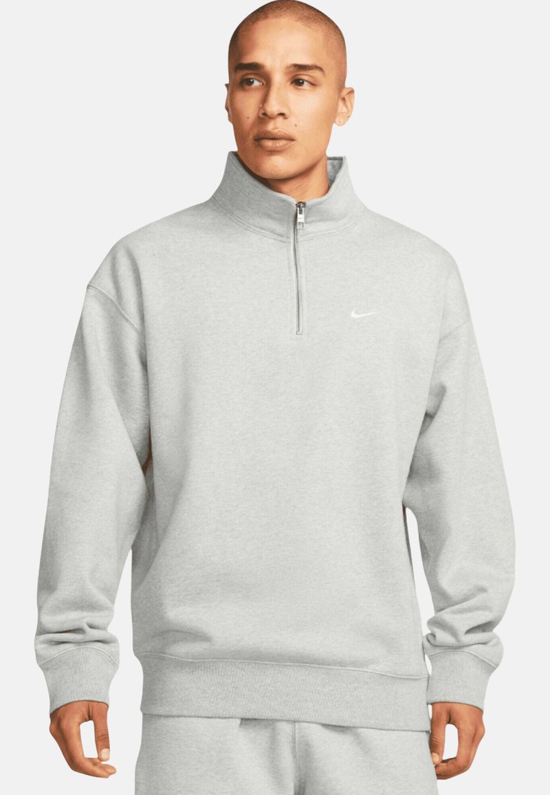 Sudadera gris de Nike con cuello de media cremallera, mangas largas, puños y dobladillo acanalados. Presenta un pequeño logo blanco en el pecho.