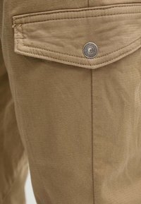 Pantalones caqui con una tela texturizada y tejida. Presenta un único bolsillo trasero con un broche de metal y detalles de costura.
