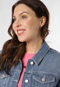 Giacca di jeans azzurro chiaro con bottoni a pressione argentati, due tasche sul petto e colletto, indossata sopra una camicia rosa con un logo.