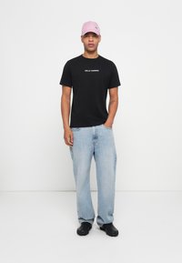 Man wearing a black Helly Hansen t-shirt, light blue baggy jeans, black shoes, and a pink Puma baseball cap, standing against a white background.