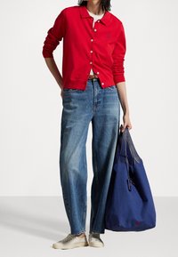 Gilet en coton rouge à boutons sur une chemise blanche, associé à un jean en denim bleu ample et un sac fourre-tout marine avec des accents en maille.