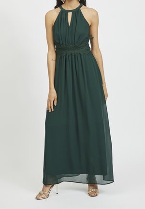 Frau trägt ein dunkelgrünes ärmelloses Maxikleid mit Neckholder, Spitzengürtel und goldenen Sandalen mit hohen Absätzen, steht vor weißem Hintergrund.