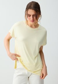 Femme aux cheveux bruns ondulés, portant des boucles d'oreilles créoles en or, un t-shirt ample jaune pâle et un pantalon blanc, tenant l'ourlet de son t-shirt.