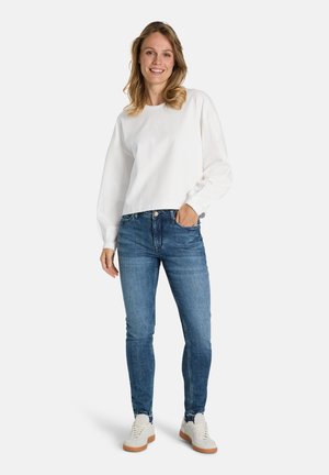 Junge Frau lächelt, trägt ein weißes Langarmshirt, blaue Jeans und weiße Turnschuhe, steht mit einer Hand in der Hosentasche.