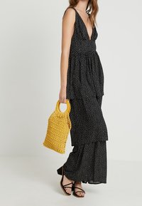 Abito maxi nero a pois con scollatura profonda e design a più strati. Abbinato a una borsa intrecciata gialla e sandali neri con cinturino.