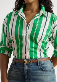 Camisa de botão às riscas verdes e brancas com gola descontraída, combinada com um cinto castanho com ferragens douradas e jeans de ganga.