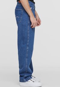 Mėlyni džinsai su laisvu siluetu, tiesiomis klešnėmis ir matomu logotipu ant užpakalinės kišenės. Derinama su baltais sportbačiais.