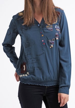 Femme portant un chemisier à manches longues bleu avec des patchs brodés colorés et des boutons, associé à un pantalon noir sur un fond blanc.