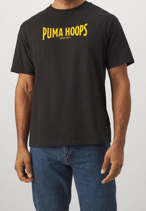 Mand iført sort t-shirt med gul tekst "PUMA HOOPS SINCE 1973" og blå jeans, stående foran en lysegrå baggrund.