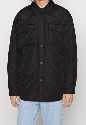 Mann trägt eine schwarze gesteppte Jacke mit Knöpfen und zwei Brusttaschen mit Klappen sowie hellblaue Jeans und steht vor einem einfarbigen Hintergrund.