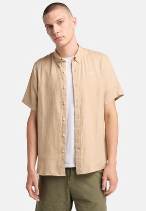 Camicia beige a maniche corte con colletto button-down, caratterizzata da un logo discreto sul petto, indossata sopra una t-shirt bianca e pantaloni verde oliva.