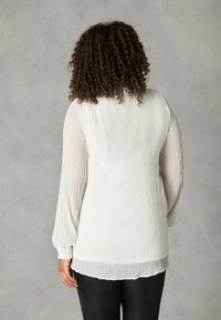 Persona dai capelli ricci che indossa una blusa bianca a maniche lunghe con texture e pantaloni neri, in piedi di spalle rispetto alla fotocamera.