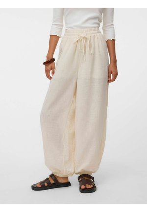 Femme portant un pantalon ample couleur crème avec une taille à cordon et des chevilles retroussées, associé à des sandales noires et un haut blanc.