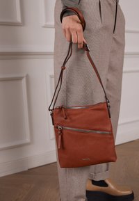 Braune Leder-Crossbody-Tasche mit zwei Reißverschlusstaschen, verstellbarem Tragegurt und weicher Textur. Ausgestellt mit karierten Hosen.