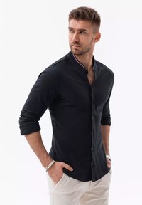 Zwarte langemouwen shirt met een staande kraag, knoopsluiting en kleurrijke afwerking. Model draagt een lichte broek en accessoires.