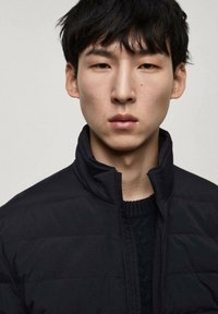 Veste matelassée noire avec un col haut et une fermeture éclair à l'avant, portée sur un pull texturé sombre. Tissu lisse avec un design épuré et minimaliste.