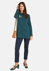 Grün bedruckte Tunika mit floralem Muster, lange Ärmel, kombiniert mit marineblauen Hosen und grünen Blockabsatzschuhen. Enthält eine kleine dekorative Brosche.