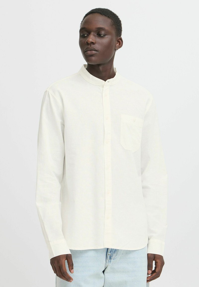 Camisa de manga larga color blanco roto con cuello alto, cierre de botones en la parte delantera y un solo bolsillo en el pecho, combinada con jeans de denim claros. Textura suave.