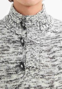 Suéter de punto con cuello alto, que presenta un patrón jaspeado en blanco y negro y tres botones de toggle negros en la parte frontal.