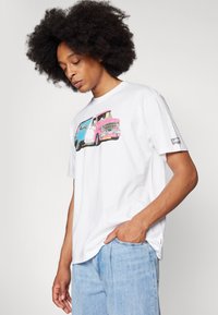 Tricou alb din bumbac cu un grafic colorat al unei furgonete vintage. Mâneci scurte, croială lejeră, cu etichetă Levi's pe partea stângă. Blugi din denim blue deschis.