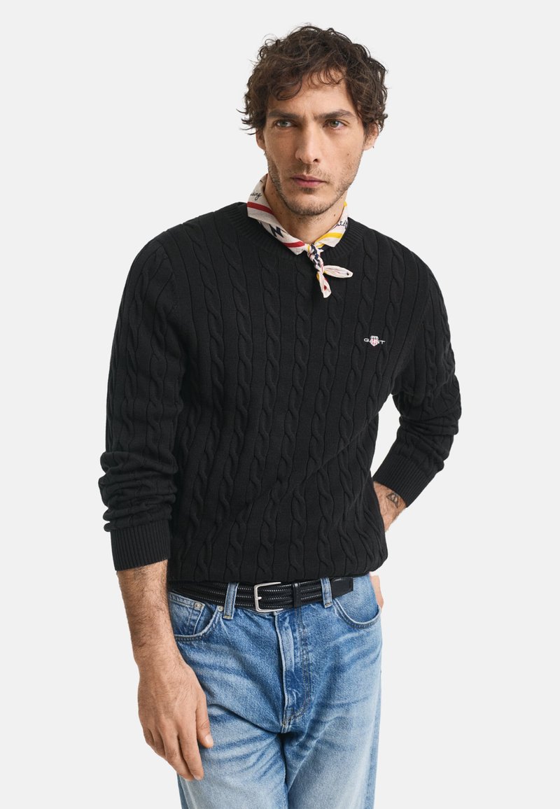 Pulover negru cu tricotaj în stil cablu, cu guler rotund, purtat peste o eșarfă cu model, asociat cu blugi de denim albastru deschis și o curea neagră.