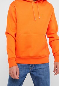 Orange hoodie i bomullsmix, med en känguruficka, dragkedjehuva och ribbade muddar. Enkel broderad detalj.