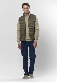 Kevyt oliivinvihreä toppaliivi beigen fleece-pulloverin päällä, yhdistetty tummansinisiin farkkuihin ja vaaleanruskeisiin tennareihin. Yksinkertainen muotoilu, ei näkyviä logoja.