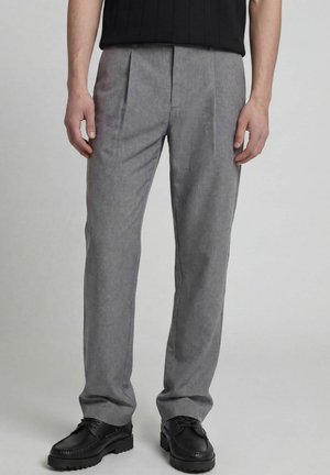 Homme portant un pantalon gris formel et des chaussures noires, debout devant un fond clair uni.