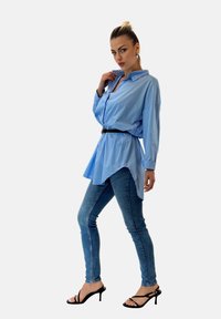 Camicia oversize azzurro chiaro con colletto a punta, cintrata in vita con una cintura nera, abbinata a jeans denim aderenti e sandali neri con tacco.