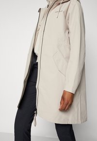 Veste imperméable beige avec une fermeture éclair à l'avant, une capuche réglable, des poches latérales et des poignets élastiques. Fabriquée en matériau résistant à l'eau.