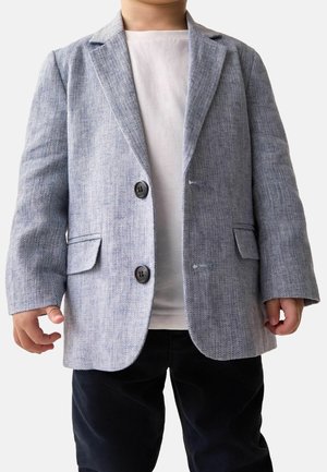Lichtgrijs, gestructureerd colbert met twee knopen en voorzakken, gedragen over een wit overhemd. Gecombineerd met een donkere broek.