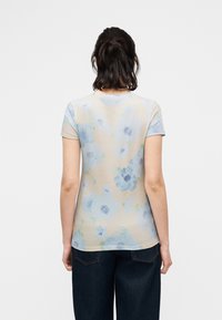 Femme aux cheveux foncés portant un t-shirt beige à motifs floraux bleu clair et un jean bleu foncé, vue de dos sur un fond uni.