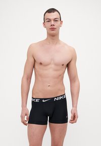 Czarne bokserki Nike z szerokim brandowanym pasem, o gładkiej fakturze i kontrastujących białych akcentach z logo. Model stoi w pozycji wyprostowanej.