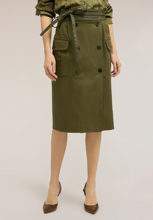 Donna che indossa una gonna verde oliva al ginocchio con dettagli a bottoni, tasche con pattina e cintura annodata, abbinata a décolleté marroni con punta appuntita.