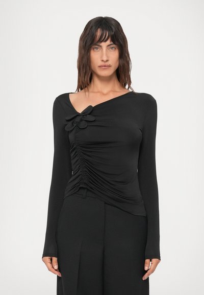 Donna con capelli di lunghezza spalla che indossa un top nero aderente a maniche lunghe con dettagli arricciati e due fiocchi in tessuto sul petto.