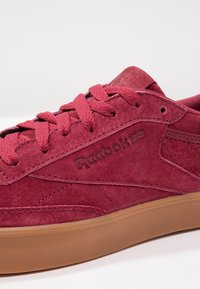 Gros plan d'une basket Reebok en daim rouge avec des lacets assortis et une semelle en caoutchouc marron sur fond blanc.