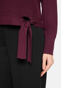 Bordeauxrode trui met een zijstrikdetail, gemaakt van een gestructureerd gebreide stof. Draagt over een zwarte broek en toont een moderne, aansluitende pasvorm.