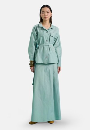Donna che indossa una giacca verde chiaro con cintura e bottoni e una lunga gonna abbinata, accessoriata con grandi orecchini e molteplici bracciali.
