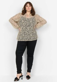 Blonde bluse i beige og sort med puffede lange ærmer, rund halsudskæring og intrikate blomster mønstre. Sammen med sorte tætsiddende bukser og højhælede sko.