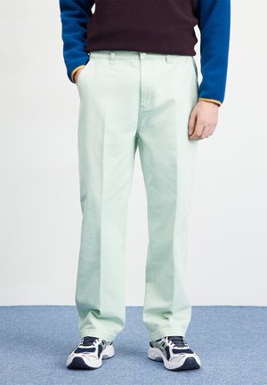 Light green, straight-leg trousers with a front pleat, made of cotton. Paired with navy and blue pullover and dark athletic shoes.