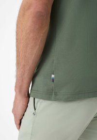 Camicetta verde a maniche corte con una texture liscia e un piccolo etichetta multicolore sul lato. Abbinata a pantaloni verde chiaro.