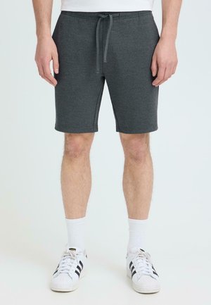 Hombre con pantalones cortos de cordón de color gris oscuro, calcetines blancos hasta la mitad de la pantorrilla y zapatillas blancas con rayas negras, posando frente a un fondo neutro.