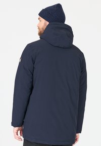 Marinblå isolerad jacka med huva, med en slät, matt yta och en logotyp på överarmen. En stickad mössa i ull bärs.