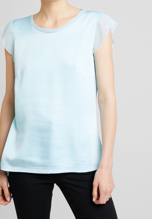 Person trägt ein hellblaues kurzärmeliges Oberteil mit Rüschenärmeln und schwarze Hose, steht vor einem einfarbigen grauen Hintergrund.