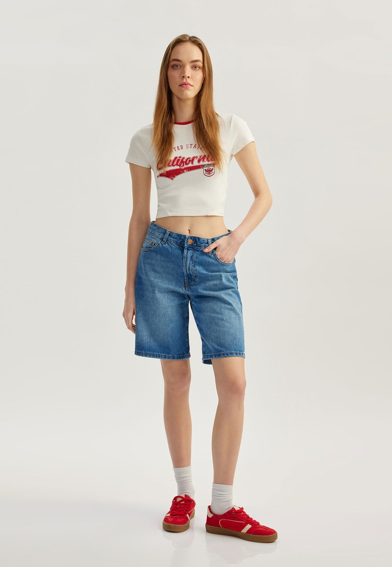 Camiseta blanca cortada con texto gráfico rojo, combinada con pantalones cortos de mezclilla azul. El modelo lleva zapatillas rojas y calcetas blancas. Tela suave, ajuste casual.