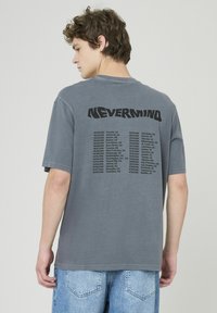 T-shirt en coton gris avec un imprimé graphique noir dans le dos, portant le mot "NEVERMIND" et une liste de dates et de lieux en dessous.
