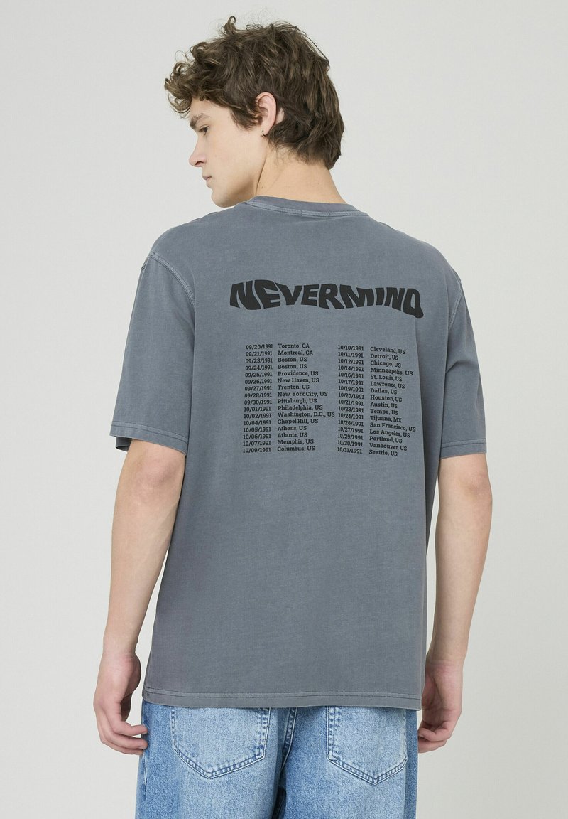 T-shirt en coton gris avec un imprimé graphique noir dans le dos, portant le mot "NEVERMIND" et une liste de dates et de lieux en dessous.