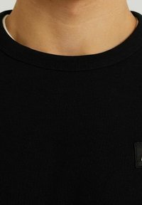 Svart bomulls-t-shirt med rund hals som har en ribbad textur och en liten svart logotypapplikation på framsidan.