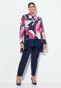 Blouse à manches longues avec un col montant, arborant un motif abstrait multicolore, associée à un pantalon bleu marine et des talons assortis.