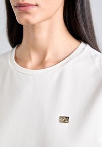 Jasno beżowa koszulka z okrągłym dekoltem o miękkiej fakturze i krótkich rękawach, z małym metalicznym logo na przodzie.